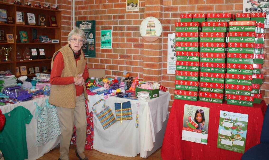 Joan Forrester stands beside the donated goodies to be packed into the 80 Operation Christmas Child Boxes stacked on a the nearby table decorated with a red cover.